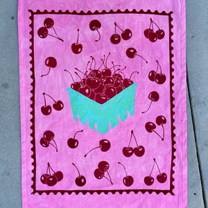 May include: A rectangular pink tea towel featuring a repeating pattern of dark red cherries and a central illustration of a light blue berry basket filled with dark red cherries, all enclosed within a dark red scalloped rectangular border.