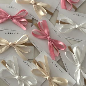May include: A collection of hair bows attached to tan elastic headbands, presented on white display cards labelled with the text ribbanie. The bows are made of smooth satin ribbon in shades of cream, white, and soft pink, featuring a knotted centre.