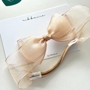 May include: A beige sheer organza bow attached to a matching elastic headband. The bow is displayed on a white card featuring the text ribbonie and ribbonie.au, with a separate dark green heart-shaped tag displaying the text ribbonie.