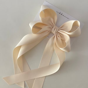 May include: A large, cream-colored satin bow with long tails. The bow is tied with a knot in the center and the tails are tied together at the bottom. The bow is on a white background with the word "ribbonie" written in black.