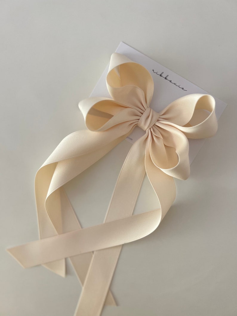 May include: A large, cream-colored satin bow with long tails. The bow is tied with a knot in the center and the tails are tied together at the bottom. The bow is on a white background with the word "ribbonie" written in black.