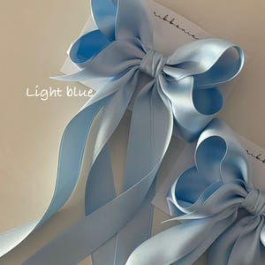 May include: Two light blue satin ribbon bows with long tails. The bows are tied together and the tails are arranged in a decorative way. The text "light blue" is written on the card below the bows.
