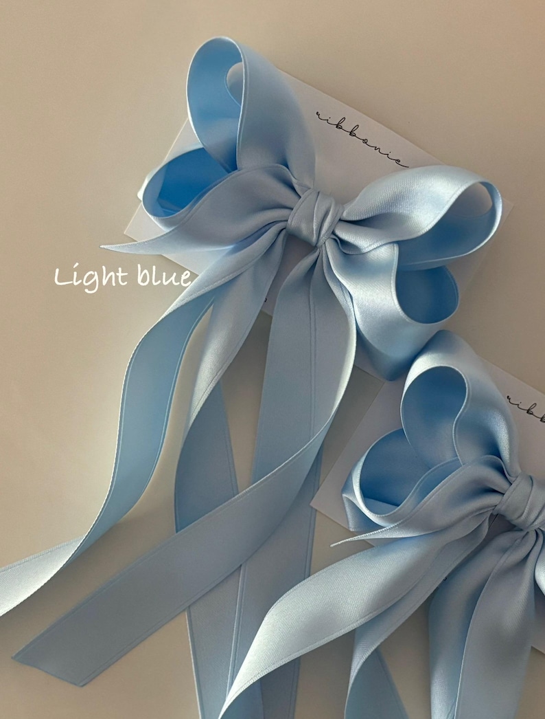 May include: Two light blue satin ribbon bows with long tails. The bows are tied together and the tails are arranged in a decorative way. The text "light blue" is written on the card below the bows.