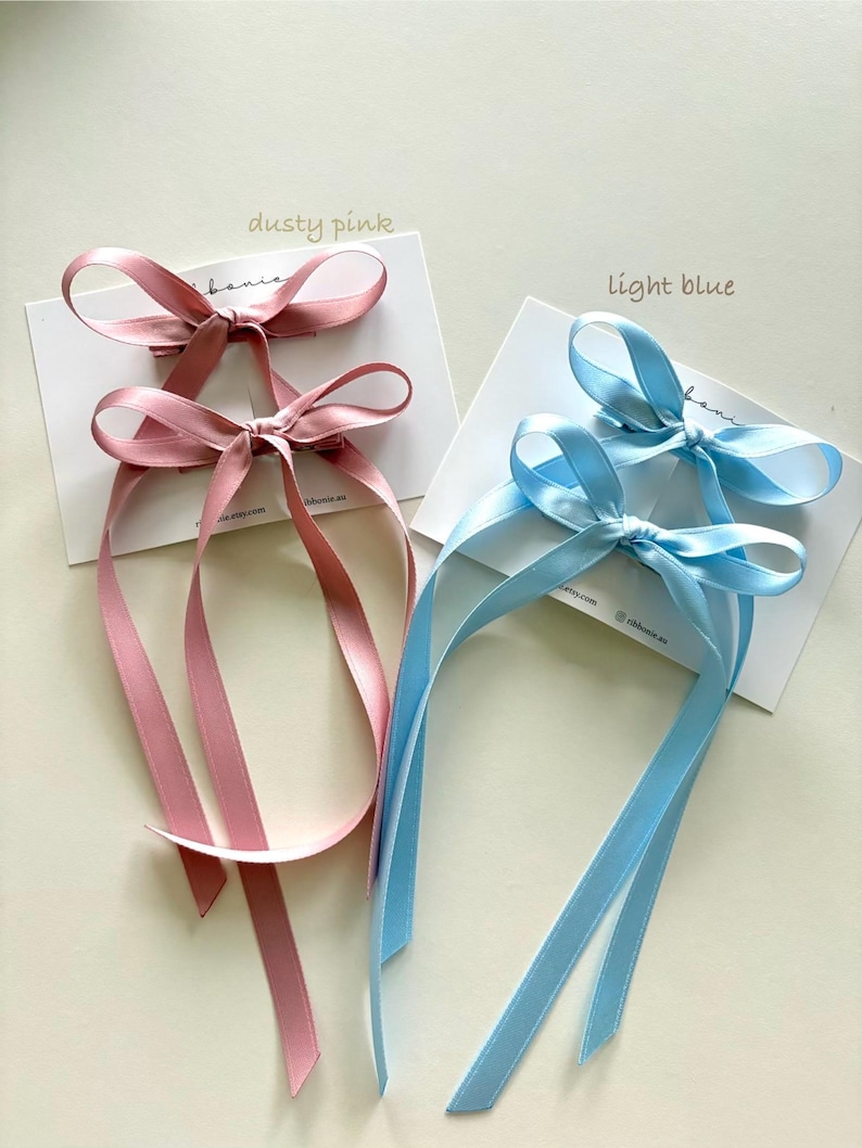 May include: Two sets of hair clips featuring satin ribbon bows, one set in dusty pink and one set in light blue. Each set is attached to a white display card. The dusty pink card is labeled dusty pink, and the light blue card is labeled light blue.