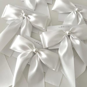 May include: A collection of white satin hair bows, each featuring a central faux pearl accent. The bows are attached to white display cards printed with the text ribbania. The smooth, reflective fabric creates a soft sheen across the surface.