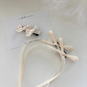Könnte beinhalten: Zwei elfenbeinfarbene Satinbänder mit schwarzen Klammern und einem langen elfenbeinfarbenen Satinband. Das Band hat einen dezenten Glanz und ist in der Mitte gefaltet. Das Band liegt auf einer weißen Oberfläche.