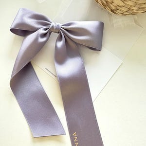 以下が含まれることがあります： 長いリボンが垂れ下がった、ラベンダー色のサテンリボン。片方のリボンには「SIENNA」の文字が金色でプリントされています。リボンは中央で結ばれており、白い表面に置かれています。
