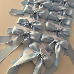 May include: A collection of light blue satin bows tied with ribbon. The bows are arranged in rows on a white surface. Each bow has a small white tag attached with the word "ribbon" printed on it.