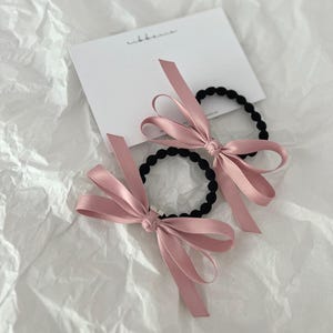 May include: Two black hair ties with pink satin bows. The bows are tied in the center and have long, flowing ribbons. A white card with the word "ribbonce" is in the background.