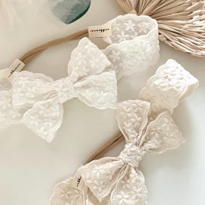 以下が含まれることがあります： 花柄刺繍のリボンが付いた、アイボリー色のベビー用ヘアバンド3つ。各ヘアバンドには、細いニュートラルカラーのバンドとブランド名が書かれた小さなタグが付いています。ヘアバンドは白い表面に配置され、背景には乾燥したヤシの葉があります。