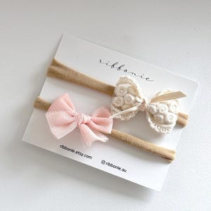May include: Two elastic headbands on a white display card labelled ribbonie, ribbonie.etsy.com, and @ribbonie.au. One headband features a light pink tulle bow, while the other displays a cream-coloured crocheted bow with a floral pattern.