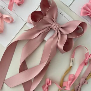 May include: A large dusty rose satin bow with long tails. The bow is tied with a knot in the center and the tails are flowing down. There are also several smaller bows in pink and dusty rose on hair elastics.