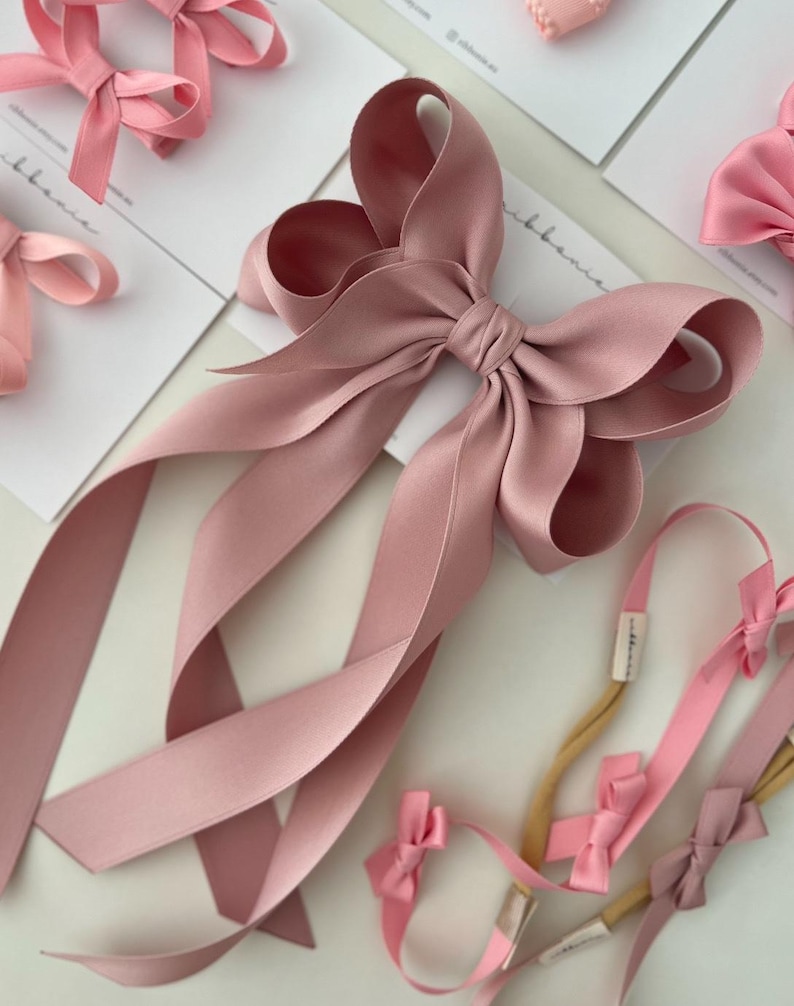 May include: A large dusty rose satin bow with long tails. The bow is tied with a knot in the center and the tails are flowing down. There are also several smaller bows in pink and dusty rose on hair elastics.