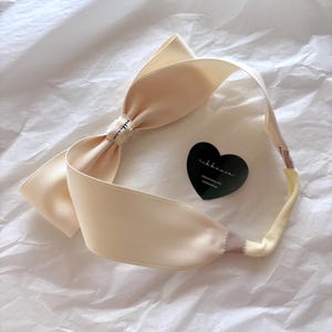 Puede incluir: Una diadema de satén color crema con un lazo grande. La diadema tiene una etiqueta en forma de corazón con el texto "libberie". La diadema está sobre una superficie blanca texturizada.