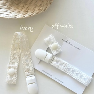 Puede incluir: Dos clips para chupete con correas de encaje blanco con patrón de corazones y clips de plástico blanco. Uno está etiquetado como ivory y el otro como off white. El clip off white está sujeto a una tarjeta blanca con el texto ribbonie y ribbonie.etsy.com.