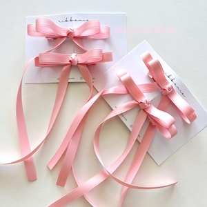 Puede incluir: Cuatro pinzas para el cabello de cinta de satén rosa claro sujetas a dos tarjetas de presentación blancas. Cada pinza presenta un lazo con dos cintas largas que cuelgan. Las tarjetas tienen impreso el nombre de marca ribbonie en cursiva negra.