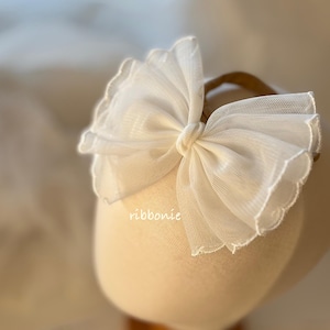 May include: A white tulle bow headband with a scalloped edge. The bow is centered on a brown headband. The word "ribbonie" is printed on the side of the headband.