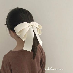 Puede incluir: Un lazo grande de satén blanco roto, accesorio para el cabello. El lazo está atado de forma clásica y se lleva en el pelo. La imagen muestra la espalda de una persona que lleva un vestido marrón. La palabra "ribbonie" está escrita en cursiva en la esquina inferior derecha.