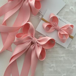 May include: Three pink satin ribbon bows of different sizes. The largest bow has long tails, the medium bow has shorter tails, and the smallest bow is attached to a wooden hair clip.