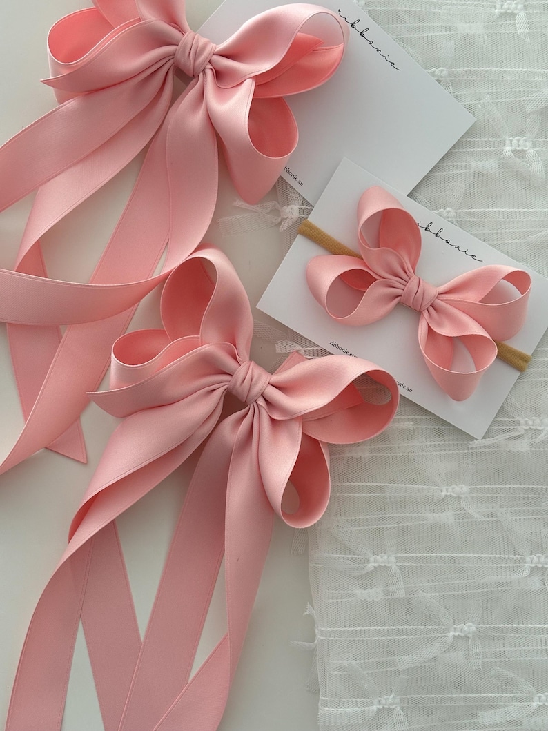 May include: Three pink satin ribbon bows of different sizes. The largest bow has long tails, the medium bow has shorter tails, and the smallest bow is attached to a wooden hair clip.