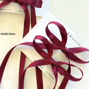 May include: Two pairs of maroon satin ribbon hair bows attached to white display cards. Each bow features a central knot and two long, trailing ribbon tails. The text "ribbonie.etsy.com" and "ribbonie.au" is printed on the cards, with "maroon" written nearby.