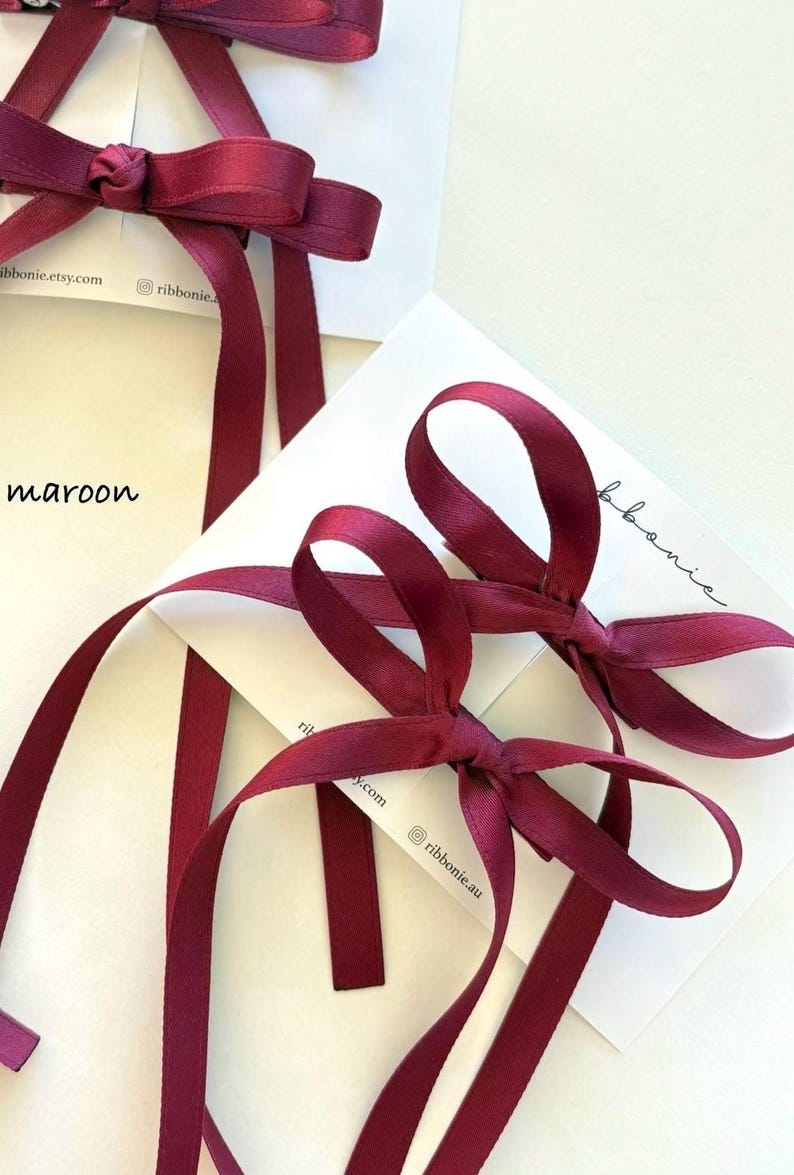May include: Two pairs of maroon satin ribbon hair bows attached to white display cards. Each bow features a central knot and two long, trailing ribbon tails. The text "ribbonie.etsy.com" and "ribbonie.au" is printed on the cards, with "maroon" written nearby.