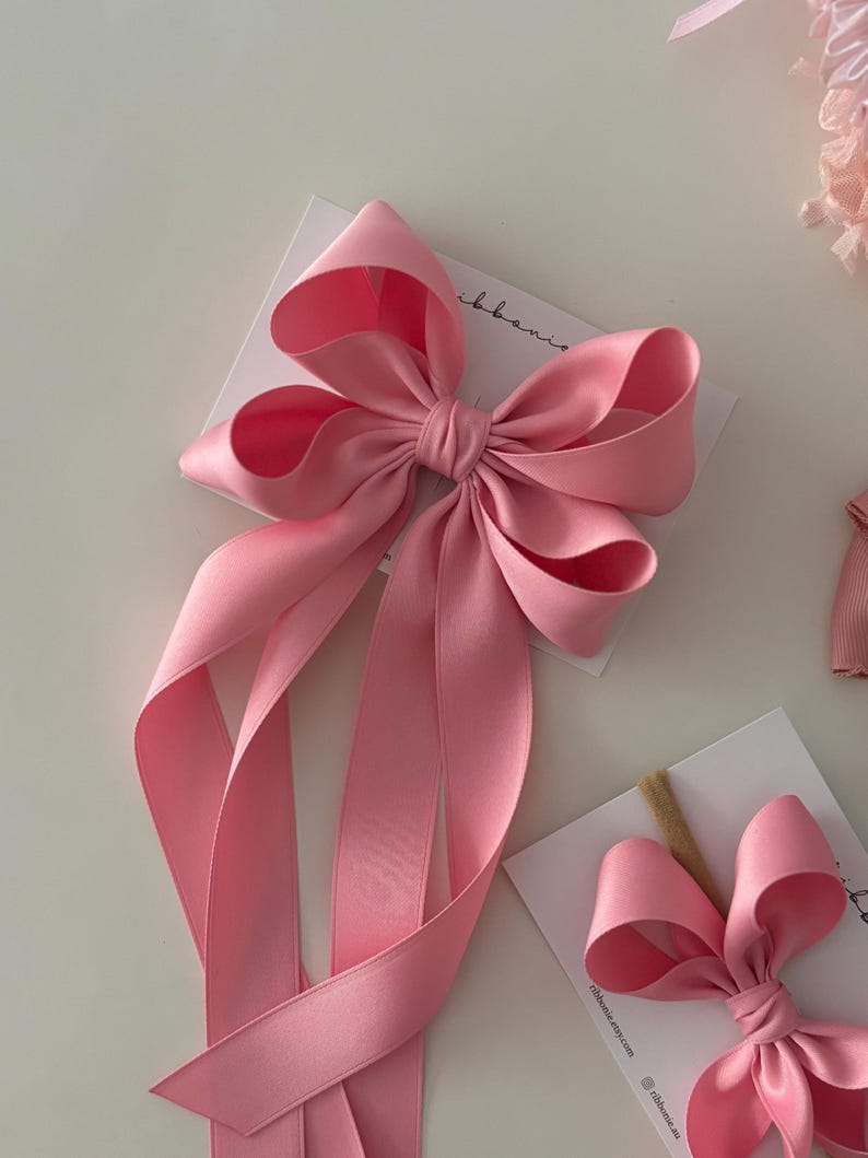 May include: A large pink satin bow with two long tails. The bow is tied with a knot in the center and the tails are flowing down. The bow is on a white background.