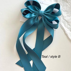 May include: Teal satin ribbon bow with long tails. The bow is tied in a simple style with two loops and a center knot. The tails are long and flowing, and the bow is perfect for adding a touch of elegance to any outfit. Teal / style B