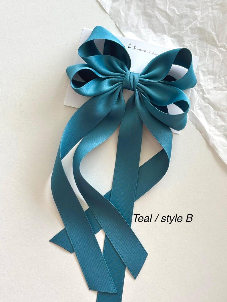 May include: Teal satin ribbon bow with long tails. The bow is tied in a simple style with two loops and a center knot. The tails are long and flowing, and the bow is perfect for adding a touch of elegance to any outfit. Teal / style B
