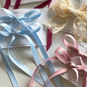 May include: A collection of hair bows on display cards, including two light blue satin bows with long tails, two dusty pink satin bows with long tails, one cream lace bow with floral embroidery, and a deep magenta satin bow with long tails.
