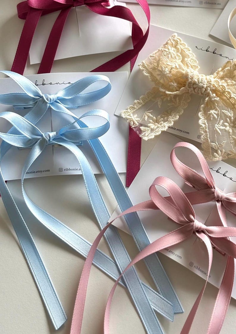 May include: A collection of hair bows on display cards, including two light blue satin bows with long tails, two dusty pink satin bows with long tails, one cream lace bow with floral embroidery, and a deep magenta satin bow with long tails.