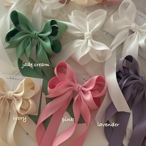 May include: Five satin ribbon hair bows in different colors: jade green, ivory, pink, lavender, and white. Each bow has long tails and is tied in a classic bow shape.