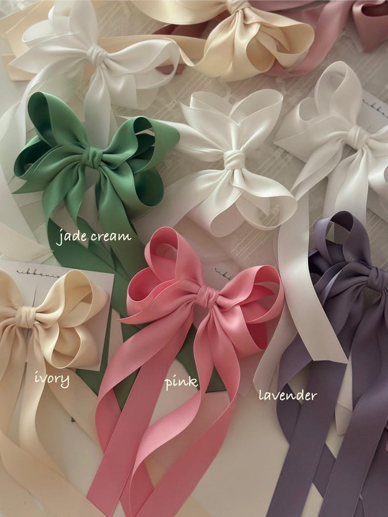 May include: Five satin ribbon hair bows in different colors: jade green, ivory, pink, lavender, and white. Each bow has long tails and is tied in a classic bow shape.