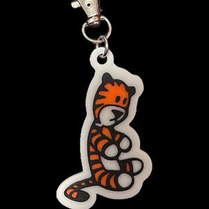 May include: A white keychain featuring a cartoon tiger design. The tiger is orange and black with a white background. The keychain has a silver metal clip.