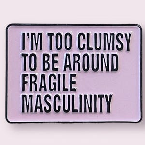 Peut inclure: Une épingle en émail rose avec une bordure noire et le texte "Je suis trop maladroit pour être autour de la masculinité fragile."