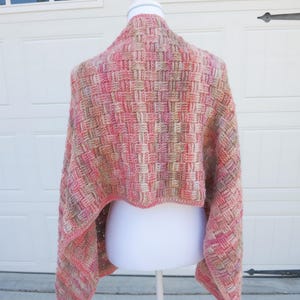 May include: A woven shawl in shades of pink, peach, and beige is displayed on a white mannequin. The shawl has a basketweave pattern and is draped over the shoulders. The background features a white garage door and a glimpse of trees.