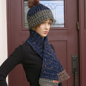 May include: A navy blue and grey crochet hat with a brown pom-pom, and a matching scarf. The scarf and hat are made from speckled yarn. The set is handmade.