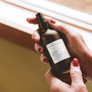 May include: A black glass bottle with a spray nozzle, labeled "Santal Beach Room & Linen Spray" by "Memory Box Candle Co."