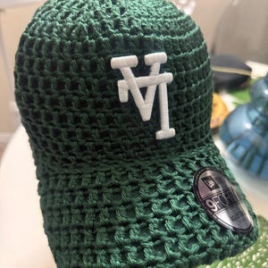 May include: A dark green crochet baseball cap featuring a white embroidered LA logo on the front. The cap has a structured crown and a curved brim, with a circular black and silver 9FORTY adjustable sticker applied to the right side of the brim.