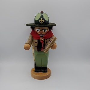 May include: A wooden nutcracker figurine of a scout, wearing a green hat with a fleur-de-lis, a red scarf, and a tan uniform. The nutcracker holds a small wooden book and stands on a wooden base.