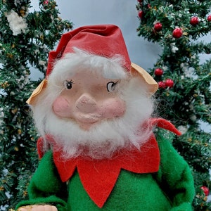 May include: A vintage Christmas elf doll with a red hat, green and red outfit, and a white beard. The elf is holding a set of gold bells.