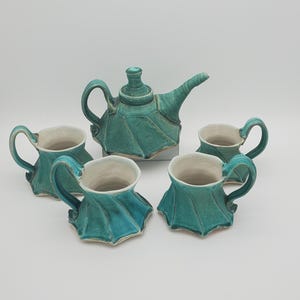 May include: A teal ceramic tea set featuring a teapot and four matching cups. The teapot has a rounded body and a curved spout. The cups have a unique, flared design. The set is a striking example of handmade pottery.