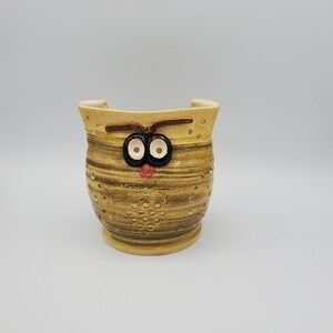 May include: A brown and yellow ceramic owl planter with black eyes and a red nose. The planter has a hole pattern on the front and sides.