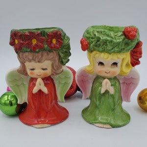 May include: Two ceramic angel candle holders with a Christmas theme. The angel on the left is wearing a red dress and a wreath of red poinsettias. The angel on the right is wearing a green dress and a wreath of green holly.