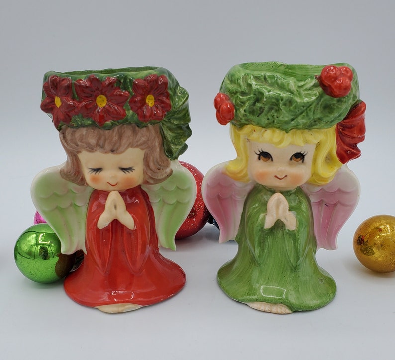 May include: Two ceramic angel candle holders with a Christmas theme. The angel on the left is wearing a red dress and a wreath of red poinsettias. The angel on the right is wearing a green dress and a wreath of green holly.