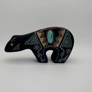 May include: A black bear figurine featuring intricate geometric patterns in turquoise, tan, and white. The design includes triangular shapes, dotted lines, and a central oval turquoise accent on the back, all set against a matte black surface.