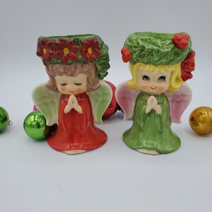 May include: Two ceramic angel candle holders with green and pink wings. The angel on the left is wearing a red dress with a green wreath of red poinsettias on her head. The angel on the right is wearing a green dress with a green wreath on her head.
