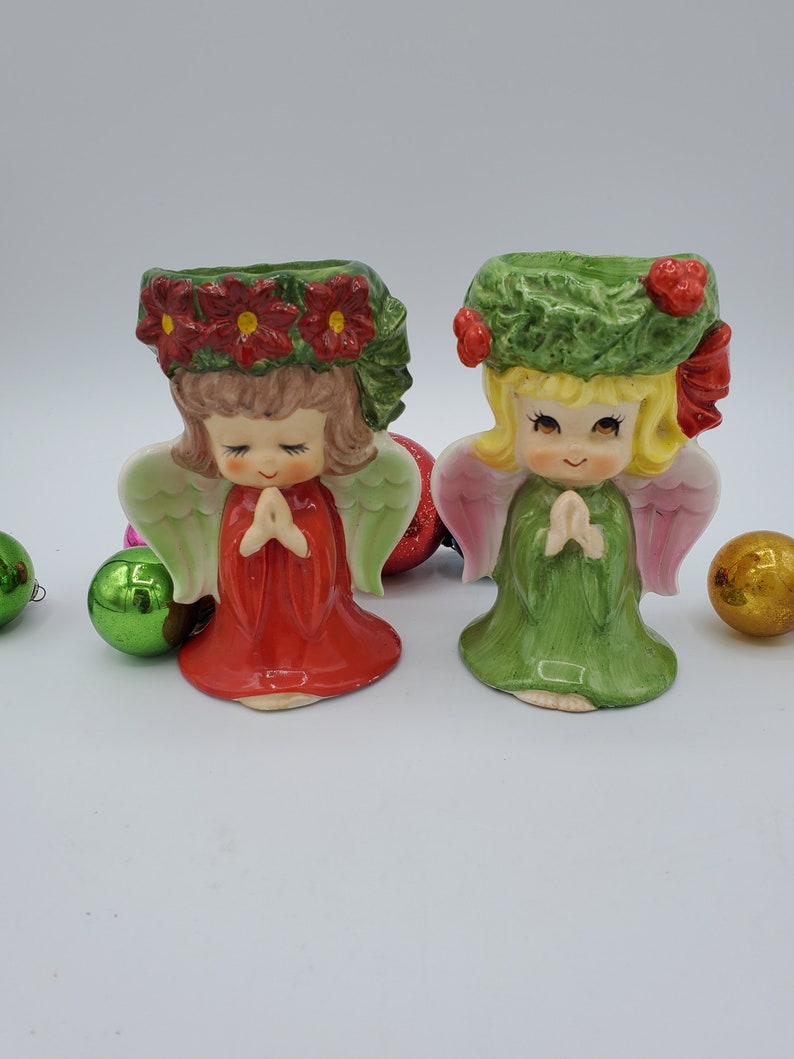 May include: Two ceramic angel candle holders with green and pink wings. The angel on the left is wearing a red dress with a green wreath of red poinsettias on her head. The angel on the right is wearing a green dress with a green wreath on her head.