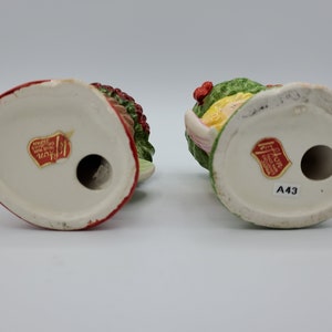 May include: Two vintage ceramic Christmas figurines, one with a red and green holly wreath and the other with a green and yellow holly wreath. Both figurines have a red sticker on the bottom with the text "Haddon Hall Hand Painted Made in Japan A43".