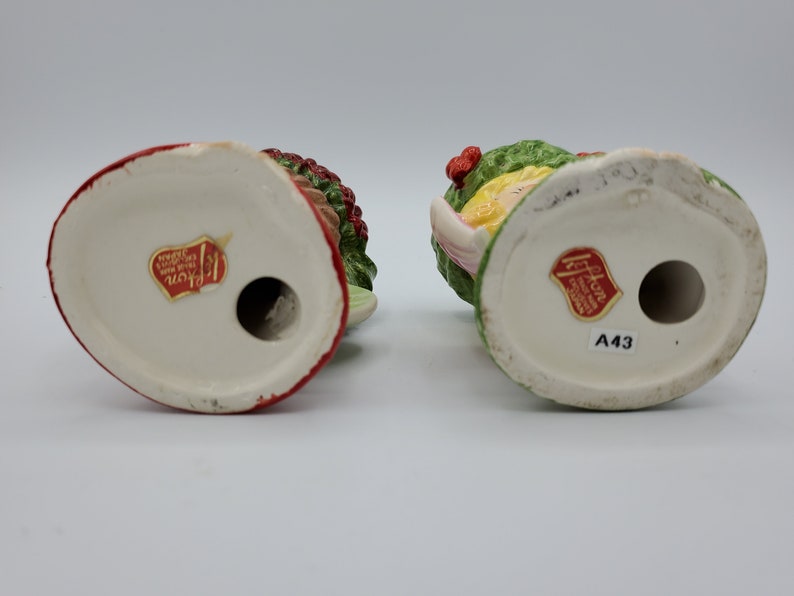 May include: Two vintage ceramic Christmas figurines, one with a red and green holly wreath and the other with a green and yellow holly wreath. Both figurines have a red sticker on the bottom with the text "Haddon Hall Hand Painted Made in Japan A43".